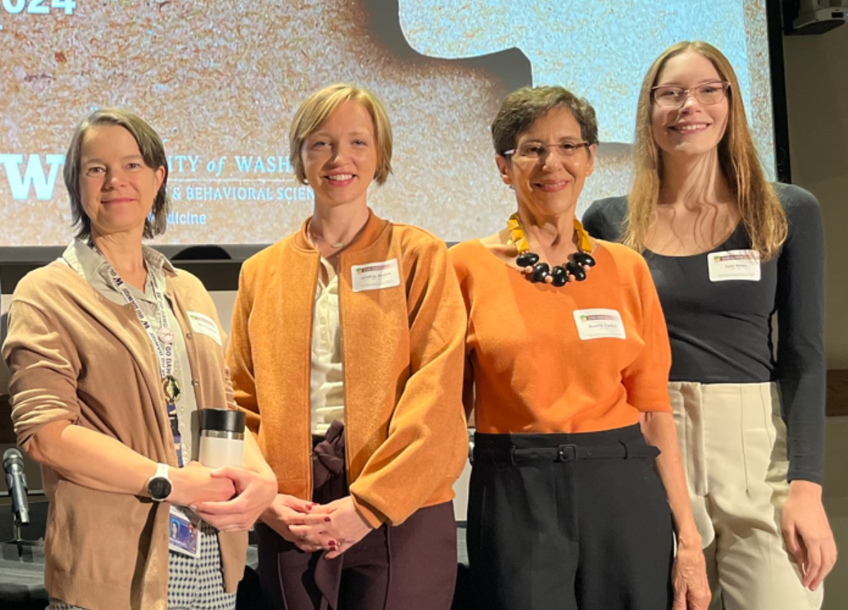 Image of four woman (named in caption) standing in a row smiling at the camera in front of a presentation screen