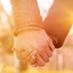 Two people's hands clasped in front of a rising or setting sun