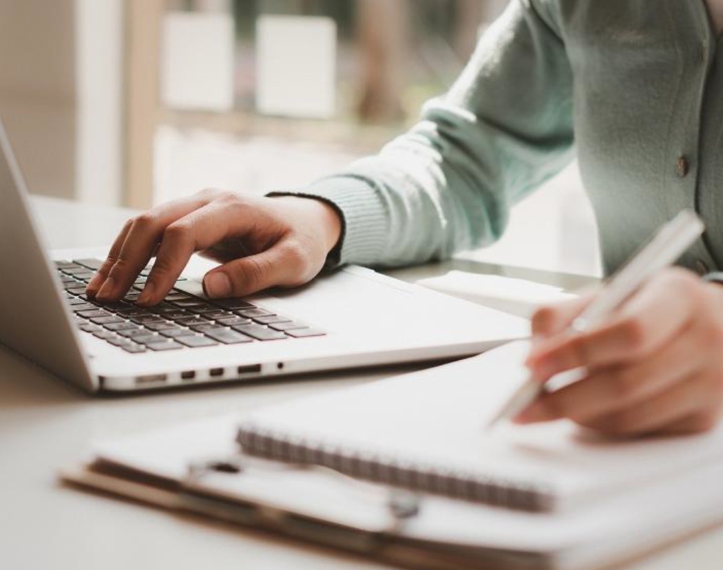 A person sitting at a laptop with one hand on the keyboard and another holding a pen at a pad of paper