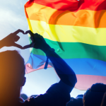 Person in shadow making a heart with their hands as a pride flag flies overhead